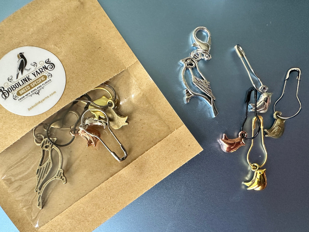 Five bird-shaped metal stitch markers in silver, rose gold, and yellow gold finishes displayed next to their kraft paper packaging on a blue surface.